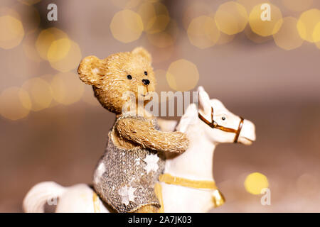 Dekorative Figuren aus einem Thema Weihnachten. Figur eines kleinen Teddybär auf einem Schaukelpferd. Weihnachtsdekoration. Festliches Dekor, warme Bokeh li Stockfoto