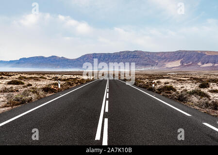 Lange, gerade, leere asphaltierten Straße durch die trockene Landschaft gegen Meer und Gebirge auf Lanzarote, Kanarische Inseln Stockfoto