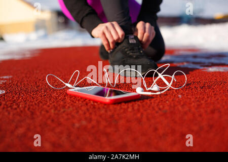 Mädchen Athlet in schwarzen Sneakers, geduckt auf der roten Spur ausgeführt werden. In der Nähe ist ein Telefon mit drahtgebundenen Kopfhörern. Kalten verschneiten Wetter. Stockfoto