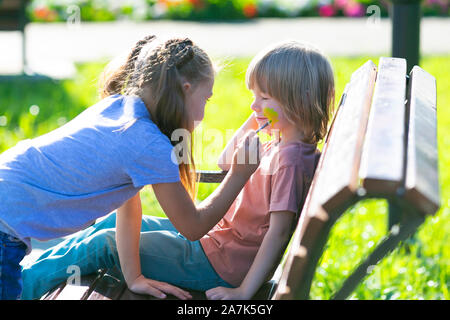 Ein kleines Mädchen ist die Anwendung aqua Make-up im Gesicht von einem 5 Jahre alten Jungen, der auf einer Bank sitzt im Park. Stockfoto