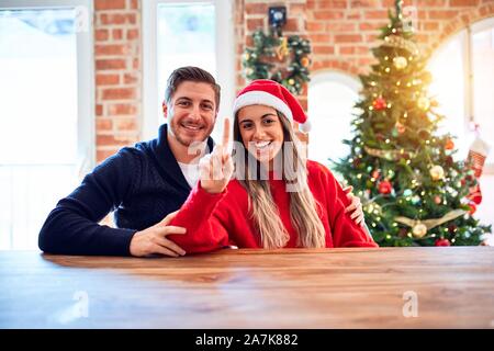 Junges Paar Tragen santa claus hat das Sitzen auf einem Stuhl und Tisch um Weihnachtsbaum zu Hause angezeigt und zeigt mit dem Finger Nummer eins Die smi Stockfoto