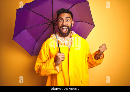 Junge indische Mann mit Regenmantel und lila Schirm über isolierte gelben Hintergrund schreien stolz und feiern Sieg und Erfolg sehr aufgeregt Stockfoto