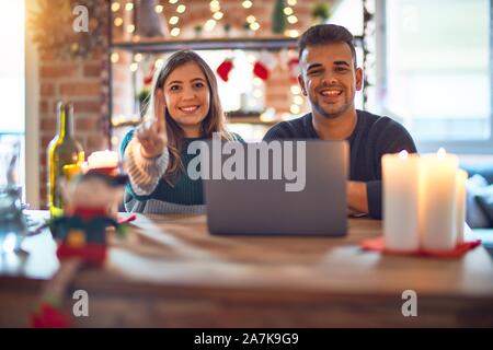Junge schöne Paar sitzt mit Laptop um Weihnachten Dekoration zu Hause angezeigt und zeigt mit dem Finger Nummer eins während lächelte zuversichtlich Stockfoto
