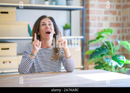 Mittleren Alters ältere Frau am Tisch sitzen zu Hause erstaunt und überrascht mit der Suche und zeigt mit Fingern und erhobenen Armen. Stockfoto