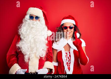 Mittleres Alter Paar Tragen santa Kostüm und Sonnenbrillen über isolierten roten Hintergrund angezeigt und zeigt mit dem Finger Nummer eins beim Lächeln konfi Stockfoto