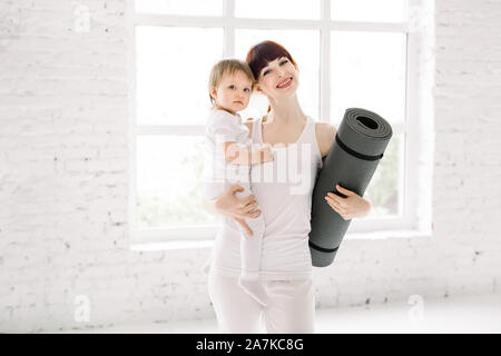 Portrait von schönen jungen Mutter in Weiß Sportbekleidung Holding ihr charmantes kleines Mädchen und Yoga Matte, Kamera und lächelnd im Stehen Stockfoto