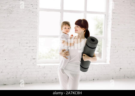 Sportlich ziemlich junge Mutter mit Baby Mädchen im weißen Sport Verschleiß und Yoga Matte in den Händen, in hellen Halle stehen. Fitness, glückliche Mutterschaft und Stockfoto