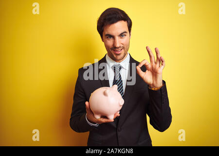 Junge gut aussehender Geschäftsmann holding Sparschwein über isolierte gelben Hintergrund tun ok Zeichen mit den Fingern, ausgezeichnete Symbol Stockfoto