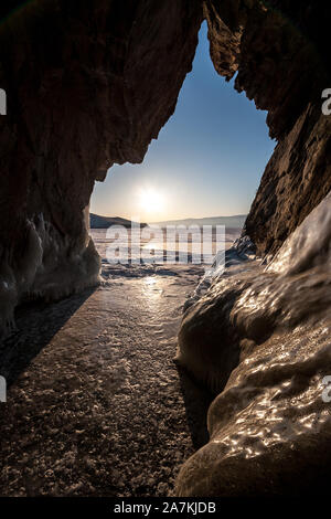 Blick aus der Höhle in den Winter am Baikalsee mit Eis bei sonnigem Wetter. Die Sonne am Horizont sichtbar ist. Es ist Eis auf dem Felsen. Vertikale Rahmen. Stockfoto