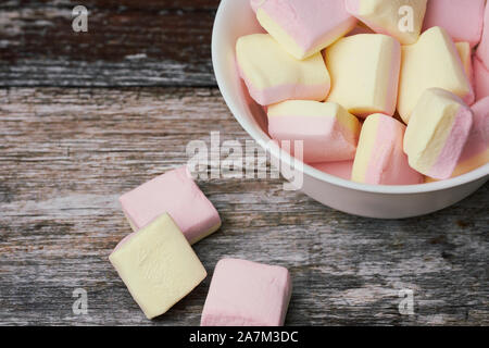 Pink und Gelb Marshmallows in einer weißen Schüssel auf Holz- Oberfläche, konzentrieren sich auf die Süßigkeiten in der Schüssel Stockfoto