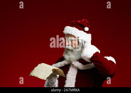 Portrait von classic Santa Leseliste auf Pergament und Anpassung der Brille beim Stehen vor rotem Hintergrund, kopieren Raum Stockfoto