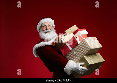 Taille bis Portrait Of Smiling Santa Claus Holding von Weihnachtsgeschenken über den roten Hintergrund stack, kopieren Raum Stockfoto
