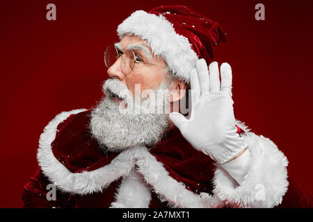 Portrait von klassischen Weihnachtsmann Hand zum Ohr versucht, Geheimnisse zu hören, während gegen den roten Hintergrund posiert im Studio Stockfoto