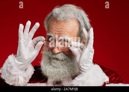 Kopf und Schultern Portrait von classic Santa Claus in die Kamera lächelnd und Anpassung der Brille, während über den roten Hintergrund posiert im Studio, kopieren Raum Stockfoto