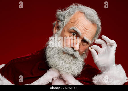 Kopf und Schultern Portrait von verdächtigen Santa Claus Anpassung der Brille und Kamera über den roten Hintergrund Suchen, Kopieren Raum Stockfoto