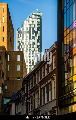 Ein schlanker, gläserner Turm – möglicherweise der 90-Meter-Beetham Tower (West Tower), erbaut 2004 – erhebt sich zwischen älteren Backstein- und Sandsteingebäuden und einer farbenfrohen Bleiglasfassade im Zentrum von Liverpool, England; zwei Flaggen wehen an einem der Gebäude im Vordergrund. Stockfoto
