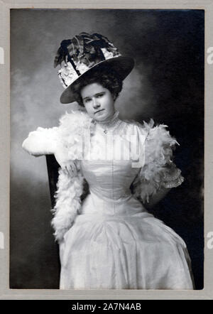 Vintage cabinet Karte zeigt eine junge Frau mit einem schicken Hut, Kleid und gefiederten Schal, ca. 1915, USA. Stockfoto