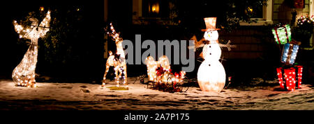 Ein Vorgarten eines Hauses ist mit hellen bis Weihnachten Dekoration in der Nacht eingerichtet. Schneemann, Schlitten, Engel und farbenfrohen Spielzeuge Boxen mit sbow unterhalb des Stockfoto