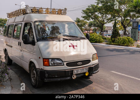 Da Nang, Vietnam - am 10. März 2019: Weiße Krankenwagen van mit rotem Kreuz auf einer breiten Straße mit grünem Laub. Stockfoto