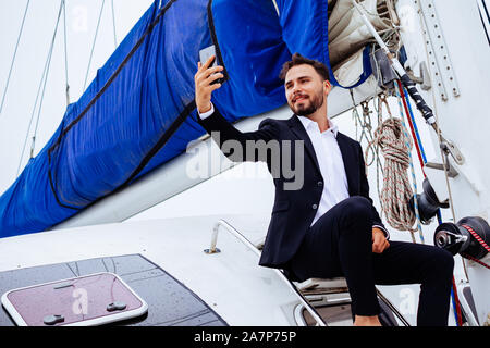 Junge schöne Geschäftsleute sitzen auf luxusyacht und unter selfies auf Handy Stockfoto
