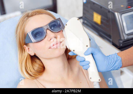 Kosmetologie Service. Junge Frau von Schönheit Klinik liegen auf medizinische Bett im Schutzbrille, blickte nachdenklich, während der Arzt mit photorejuvenation Laser Stockfoto
