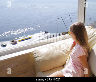 Ein kleines Mädchen sitzt am Fenster und schaut auf das Meer. Der Blick aus dem Fenster zum Meer. Rückansicht der jungen Frau an der Meerblick vom Balkon Stockfoto