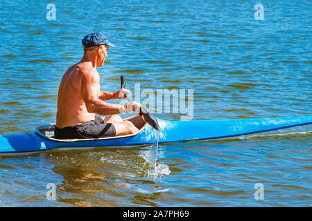 Man paddeln eine blaue und gelbe Kajak auf dem Fluss in der Nähe der Küste. Kajak Konzept. ein Mann schwimmt in einem Kanu auf dem Fluss. Ein Mann in einem Boot auf einem sonnigen Stockfoto