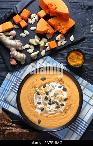 Nahaufnahme der cremige geröstete butternut Kürbis curry bestreut mit Kürbiskerne in einem schwarzen Schüssel auf einem Holztisch, Inhaltsstoffe auf einer Schiefertafel, Anzeigen Stockfoto