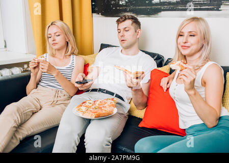 Junge müde Leute in ungezwungener Kleidung essen Pizza, sitzen auf der Couch und beobachtete, langweiligen Film im TV, Ruhe zu Hause. Wochenende, Freizeit, Fast food Konzept Stockfoto