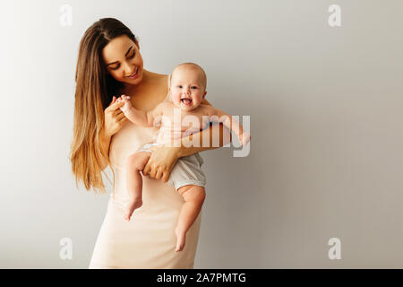 Schöne glückliche Mutter hält Ihr Baby auf einem einfachen hellen Hintergrund. Kind lächelt Stockfoto