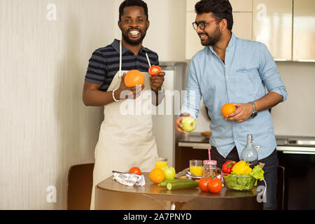 Zwei männliche Studenten in Freizeitkleidung zu kochen Smoothie in der Küche der Herberge, die hielten Grapefruit, Orange, Apfel, Tomate in die Hände gehen, Jubeln