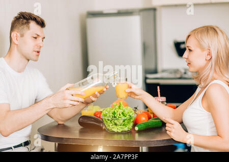 Portrait von hübschen jungen Paar in Liebe in leckeres Frühstück am Tisch in der Küche sitzt. Schöner Mann gießen Saft aus Glas für seine Frau Stockfoto