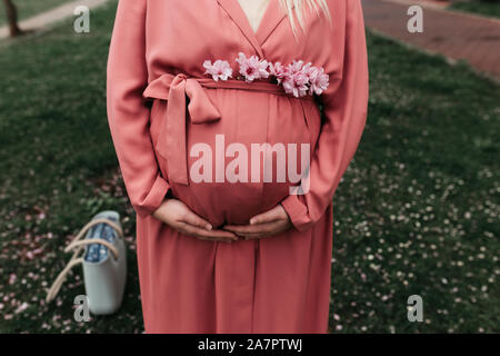 Schöne schwangere Frauen in rosa Outfit fotografiert nächsten Baum im Frühling blühen. Nahaufnahme Foto von ihrem Bauch und ohne Gesicht dargestellt. Stockfoto