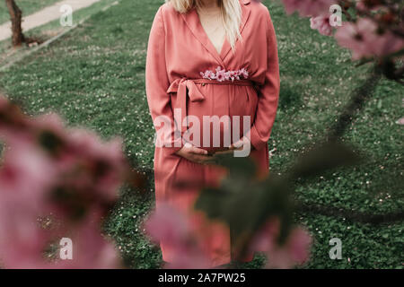 Schöne schwangere Frauen in rosa Outfit fotografiert nächsten Baum im Frühling blühen. Nahaufnahme Foto von ihrem Bauch und ohne Gesicht dargestellt. Stockfoto