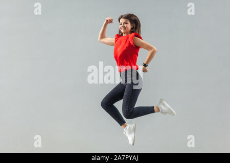 Voller länge des gerne schöne Brünette junge Frau in casual rotes Hemd springen, toothy Lächeln und Kamera mit Glück. Indoor, Studio Stockfoto