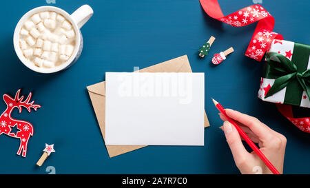 Weibliche Hand schreiben Brief an Santa Claus auf dunkelblauem Hintergrund mit Geschenke, Dekorationen und Kaffee Tasse. Draufsicht auf vintage Tabelle. Weihnachten lette Stockfoto