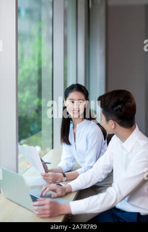 Taille bis Schuß von zwei lächelnden jungen männlichen und weiblichen Geschäftsleuten mit schwarzem Haar reden während an einem Schreibtisch in einem Büro arbeiten Stockfoto