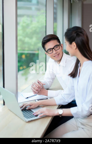 Taille bis Schuß von zwei lächelnden jungen männlichen und weiblichen Geschäftsleuten mit schwarzem Haar reden während an einem Schreibtisch in einem Büro arbeiten Stockfoto