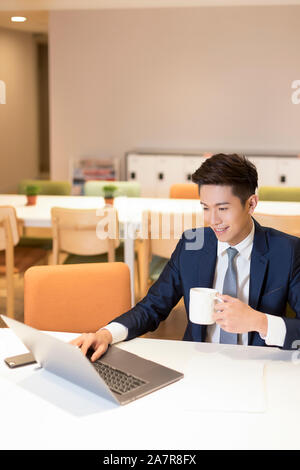 Eine lächelnde junge lächelnde Geschäftsmann im Anzug an einem Schreibtisch in einem Büro vor einem Laptop und Kaffee trinken und Arbeiten, Taiwan sitzen Stockfoto