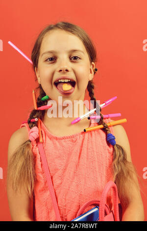 Mädchen hält Schultasche in den Händen und herzförmige Plätzchen auf der Zunge. Schule Mädchen mit glücklichen Gesicht auf Lachs Hintergrund isoliert. Zurück zum Konzept der Schule. Kind in rosa Kleid mit Zöpfen und Buntstifte. Stockfoto