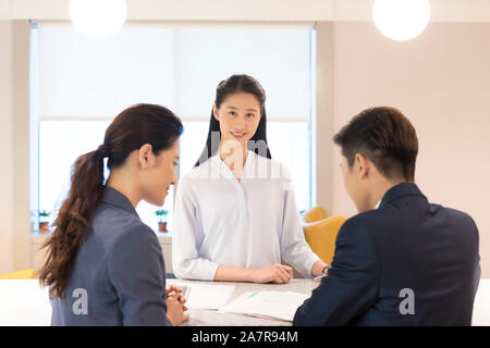 Lächelnden jungen Geschäftsfrau mit langen schwarzen Haare in die Kamera schaut mit zwei anderen Geschäftsleuten im Vordergrund sichtbar Stockfoto