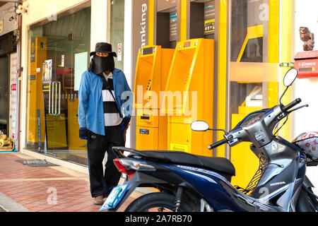 Khao Lak, Thailand, 30. August 2019: Asiatischer Mann in Cowboyhut und Stirnband in der Nähe von Geldautomaten, möglicherweise plotten Kriminalität, schaut Fotograf aussetzen. Stockfoto