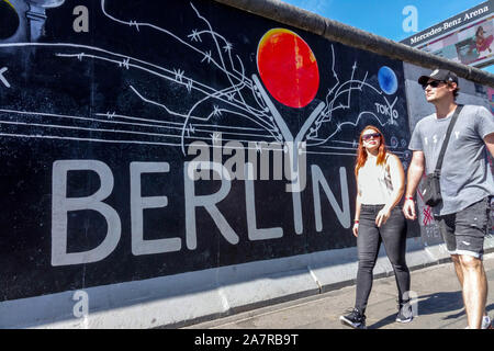Junge Menschen, die auf dem Bürgersteig neben der historischen Berliner Mauer in der East Side Gallery im Stadtteil Friedrichshain in Berlin laufen Stockfoto