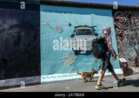 Berliner Mauer Graffiti East Side Gallery Deutschland junger Mann zu Fuß Der Hund, der um Trabant Auto Friedrichshain City Street Berlin Stadtmenschen Stockfoto