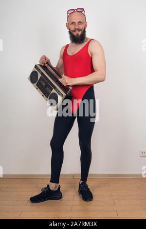 Ein bärtiger Mann in einem roten Strampler und leggings mit Sonnenbrille auf dem Kopf hält ein Tonbandgerät. Das Bild einer Retro Party im Stil der 90er Jahre. Stockfoto