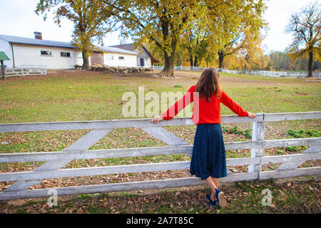 Ein schönes Mädchen in einem roten Pullover steht mit dem Rücken in der Nähe von einem weißen Holzzaun. Schönen Herbst Stockfoto