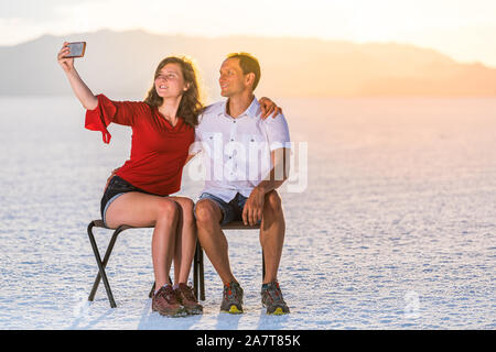 Junge romantische Paare sitzen auf Stühlen unter selfie Bild mit Telefon auf weißen Bonneville Salt Flats in der Nähe von Salt Lake City, Utah und Bergblick Stockfoto