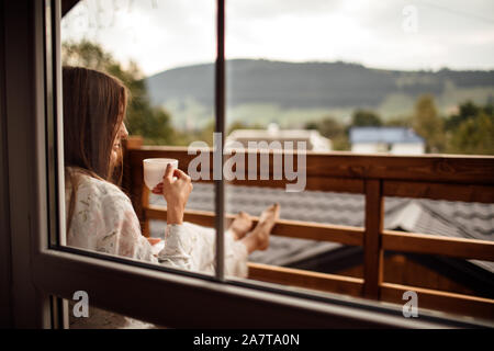 Junge Frau auf dem Balkon mit einer Tasse Kaffee oder Tee am Morgen. Sie im Hotel Zimmer an der Natur in Sumer. Mädchen ist in stylis gekleidet Stockfoto