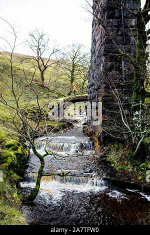 Eine Frau kreuzt eine kleine Brücke aus Stein unter einem Eisenbahnviadukt bei Cowgill, in der Nähe von Ribblehead, in Dentdale, North Yorkshire National Park. Stockfoto