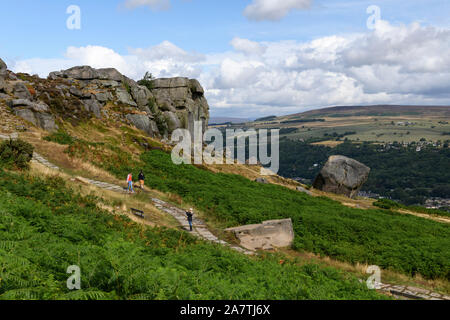 Menschen zu Fuß auf dem Weg von hohen Sonnenbeschienenen Felsvorsprung (Tal) - die malerische Landschaft von Kuh und Kalb Felsen, Ilkley Moor, West Yorkshire, England, UK. Stockfoto